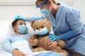 Nurse takes care of the patient child in hospital bed playing with teddy bear, wearing protective masks, corona virus covid 19