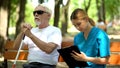 Nurse taking notes sitting by blind old patient in park, walking with disabled