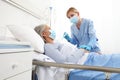 Nurse with thermometer measures fever on elderly woman patient lying in the hospital room bed, wearing protective gloves and