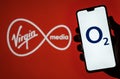 O2 Telefonica logo on smartphone screen hold in hand and Virgin Media logo on blurred background. Concept for Virgin and O2 merger