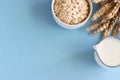 Oat milk in a jug and oats in a white ceramic bowl on a blue paper background.