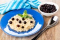 Oatmeal with milk, blueberry, leaves of mint in plate, napkin, bowl with blackcurrant, spoon on wooden table