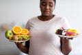 Obese Afro woman holding plates with fruits and desserts, tempted to break weight loss diet at home, selective focus