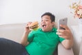 Obese person eating hamburger while using mobile phone