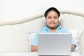 Obese schoolboy sitting and wear headphones to learn online from a laptop on their bed