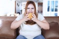 Obese woman eating burger on sofa