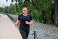 An obese young woman jogging outdoors. Fat beautiful smiling girl in a black tracksuit is engaged in fitness for weight