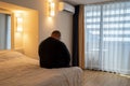 Obesity man sadly sitting on bed on vacation in hotel alone without friends having midlife crisis.