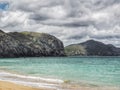 An ocean beach with rock cliffs, green hills abd cloudy sky