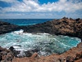 Ocean waves breaking on the rocky coast of hardened lava with caverns and cavities. Blue sky with white clouds and mountains Royalty Free Stock Photo