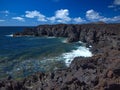 Ocean waves breaking on the rocky coast of hardened lava with caverns and cavities. Deep blue sky with white clouds and mountains Royalty Free Stock Photo