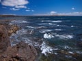 Ocean waves breaking on the rocky coast of hardened lava with caverns and cavities. Deep blue sky with white clouds and mountains Royalty Free Stock Photo