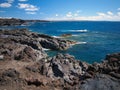 Ocean waves breaking on the rocky coast of hardened lava with caverns and cavities. Deep blue sky with white clouds and mountains Royalty Free Stock Photo