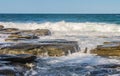 Ocean waves crash against eons old volcanic rock and water runs and breaks the stone - with tiny boats on the horizon