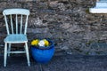 A off white painted wooden chair with a large blue ceramic plant pot with yellow and white flowers against an old stone exterior Royalty Free Stock Photo