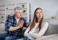 Offended mature lady sitting on couch with folded arms, her husband begging for forgiveness after fight, indoors