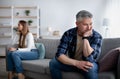 Offended mature man and woman sitting on different sides of sofa after after quarrel indoors, empty space