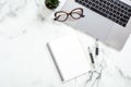 Office desk table with laptop computer, paper notepad, glasses, stationery, succulent plant on white marble surface. Flat lay, top