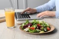 Office employee working with laptop at white wooden table, focus on fresh vegetable salad with glass of juice. Business lunch