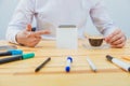 Office table with accessories: white sheet of paper, marker of blue and pink, black phone, coffee cup. With one hand