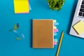 Office table desk with set of colorful supplies, white blank note pad, cup, pen, pc, crumpled paper, flower on blue Royalty Free Stock Photo