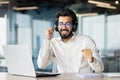 An office worker, a young Indian businessman sits in the office at the desk in headphones in front of the laptop, dances Royalty Free Stock Photo