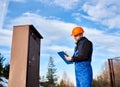 Oil inspector in protective overalls and orange helmet making notes next to a transformer