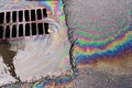 An oil slick against the backdrop of an asphalt road flows into a storm drain through a grate. Royalty Free Stock Photo