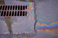 An oil slick against the backdrop of an asphalt road flows into a storm drain through a grate. Royalty Free Stock Photo