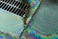 An oil slick against the backdrop of an asphalt road flows into a storm drain through a grate Royalty Free Stock Photo