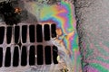 An oil slick against the backdrop of an asphalt road flows into a storm drain through a grate Royalty Free Stock Photo