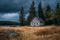 Old abandoned cabin stands in a field by a serene lake, with a stormy sky and pine trees creating a dramatic backdrop Royalty Free Stock Photo