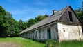 Old abandoned long house in poor condition. Bars and cracked panes in the windows. Around the tree and dandelions in the grass.