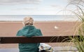 Old age - alone. Old woman with bag seen from behind sitting looking out to sea.