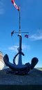 An old anchor with a ships mast in the background flying flags