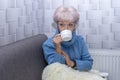 Old beautiful woman 60-65 years old drinking hot tea from a white mug, warm at the heating radiator. Concept: low indoor temperatu