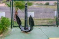 Old black anchor stands vertically close to metal green fence