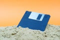 An old black floppy disk is half covered in sand