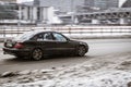 Old black Mercedes W211 on snowy winter road in city. Mercedes-Benz E-class drives on slippery snow covered street