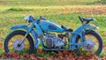 Old blue rusty Soviet motorcycle parked on green grass with fallen brown leaves