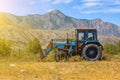 An old blue tractor excavator stands in a field with green and yellow grass against a background of mountains and blue sky during Royalty Free Stock Photo