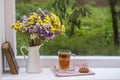 Old book, colorful bouquet of flowers and glass cup of tea on background of window with raindrops at home at summer day Royalty Free Stock Photo