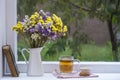 Old book, colorful bouquet of flowers and white cup of tea on background of window with raindrops at home at summer day Royalty Free Stock Photo