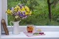 Old book, colorful bouquet of flowers and white cup of tea on background of window with raindrops at home at summer day Royalty Free Stock Photo