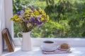 Old book, colorful bouquet of flowers and white cup of tea on background of window with raindrops at home at summer day Royalty Free Stock Photo