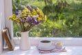 Old book, colorful bouquet of flowers and white cup of tea on background of window with raindrops at home at summer day Royalty Free Stock Photo