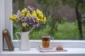 Old book, colorful bouquet of flowers and white cup of tea on background of window with raindrops at home at summer day Royalty Free Stock Photo