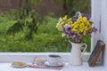 Old book, colorful bouquet of flowers and white cup of tea on background of window with raindrops at home at summer day Royalty Free Stock Photo