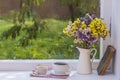 Old book, colorful bouquet of flowers and white cup of tea on background of window with raindrops at home at summer day Royalty Free Stock Photo