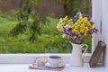 Old book, colorful bouquet of flowers and white cup of tea on background of window with raindrops at home at summer day Royalty Free Stock Photo